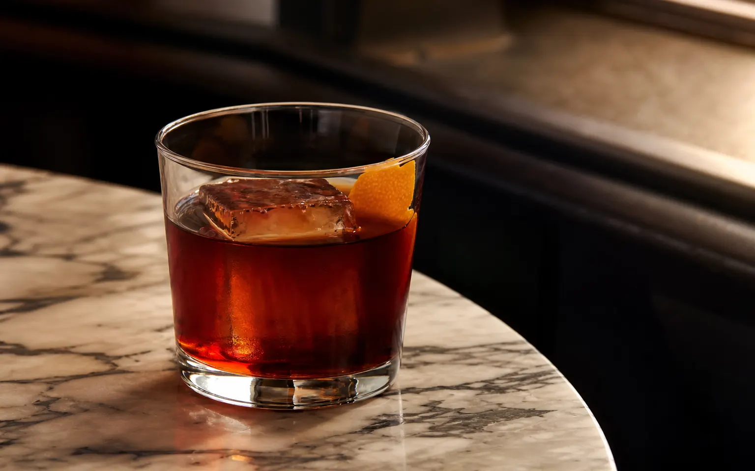 A dark red-brown Coffee Negroni over a large ice cube with orange peel