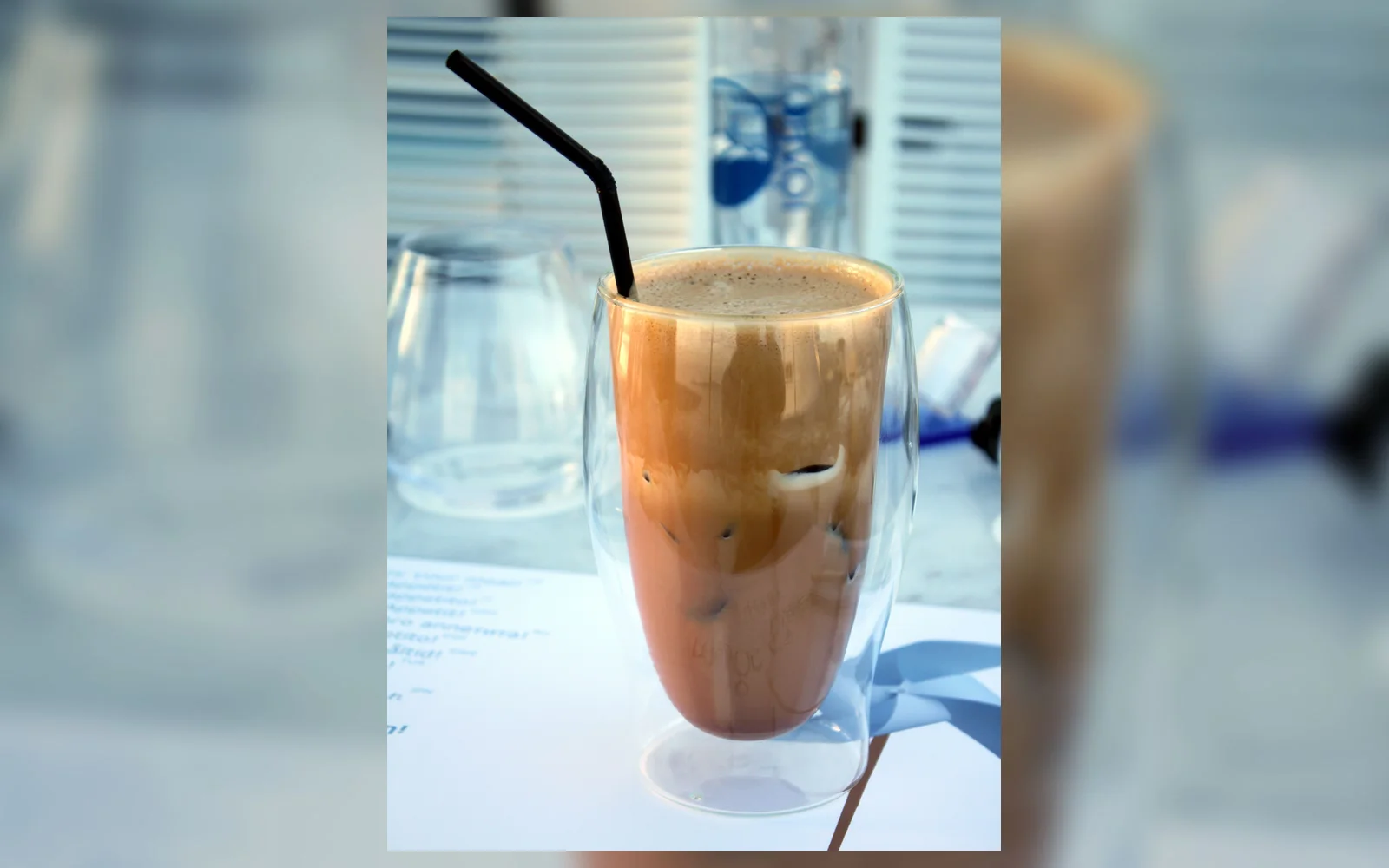 Greek frappé in a clear tall glass with ice, tan coffee foam, and a black straw
