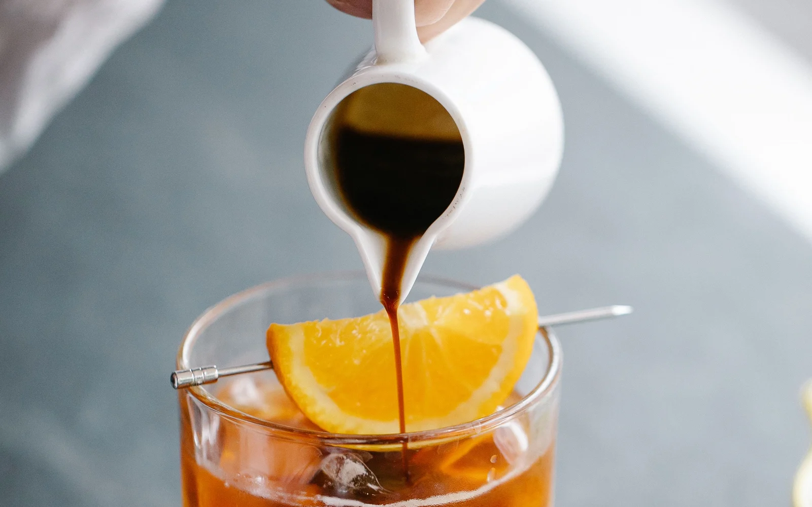 Coffee pouring from a white pitcher into an iced orange drink garnished with an orange slice